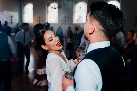 The bride, with glitter on her face, laughs next to a guest holding a glass during the party