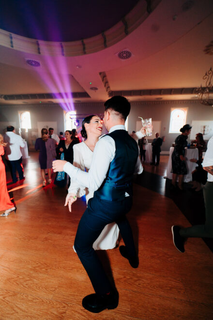 The bride dances with a guest in a blue waistcoat holding a glass, with a purple light beam in the background