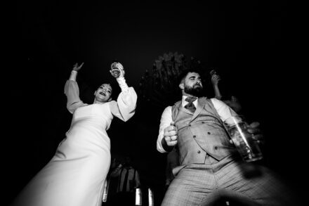 The couple dance with drinks in hand under a chandelier, seen from below in black and white