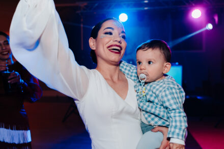 The bride, with glitter on her face, dances with one arm raised, holding the baby with a pacifier