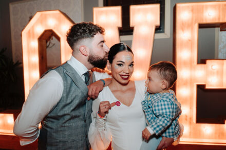 The groom kisses the bride’s head as she holds the baby in front of giant light-bulb letters