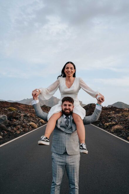 La novia sonríe sentada a hombros del novio, que la sujeta de las manos en una carretera entre campos de lava
