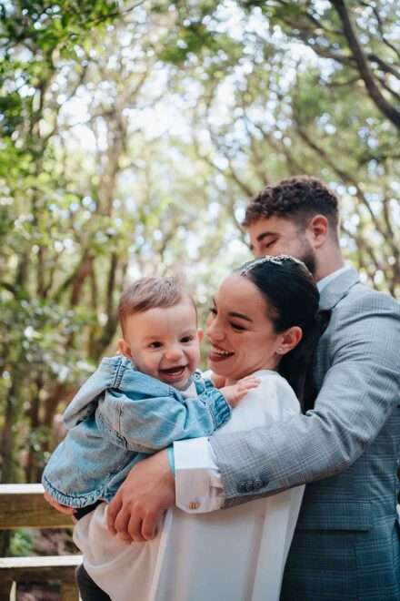 La novia sostiene en brazos a un bebé con chaqueta vaquera que ríe mientras el novio los abraza a los dos