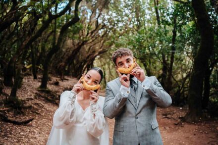 Los novios sostienen cada uno un plátano delante de la boca, como una sonrisa, en un sendero del bosque