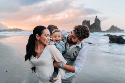 Los novios sostienen entre los dos a un bebé sonriente en la orilla, con los islotes rocosos al fondo