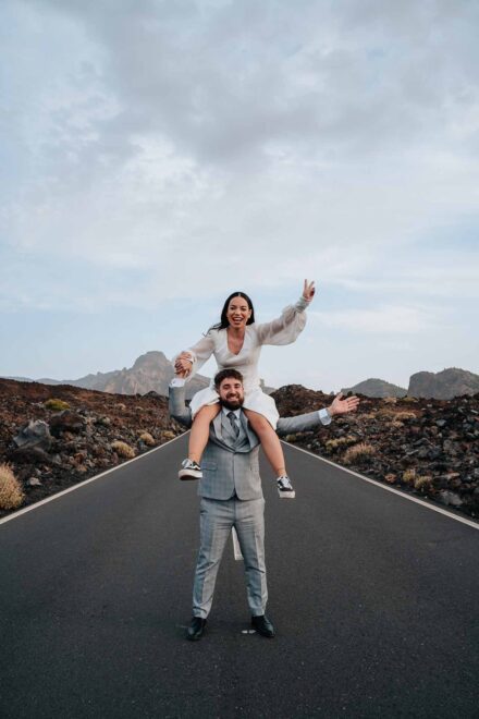La novia hace el signo de la victoria a hombros del novio, que abre los brazos en mitad de la carretera
