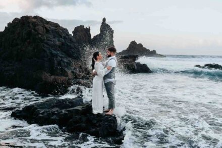 Los novios se abrazan sobre una roca negra mientras las olas rompen a su alrededor