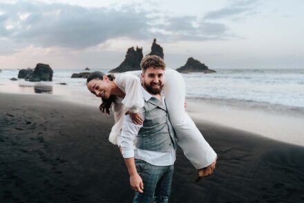 El novio carga a la novia sobre el hombro y los dos se ríen en la arena negra de la playa
