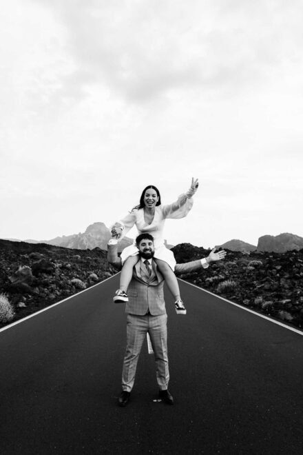 La novia a hombros del novio con la mano en alto y él con los brazos abiertos en la carretera, en blanco y negro
