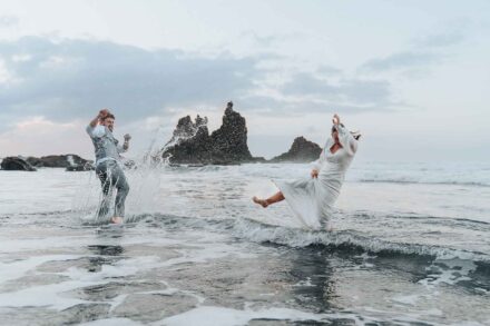 Los novios se salpican agua a patadas en la orilla, con los islotes rocosos al fondo