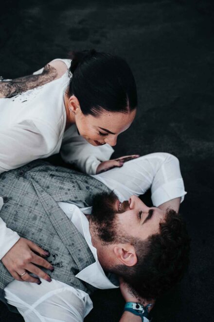 La novia, con la espalda manchada de arena, se inclina sonriendo sobre el novio tumbado en la playa