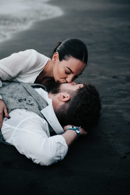 La novia besa al novio, tumbado boca arriba en la arena negra, cerca de la orilla