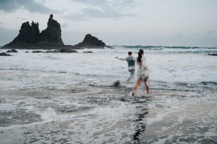 Los novios entran corriendo en el mar entre la espuma de las olas, de espaldas a la cámara