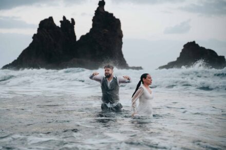 El novio sacude el pelo mojado con los brazos abiertos junto a la novia, los dos con el agua por la cintura