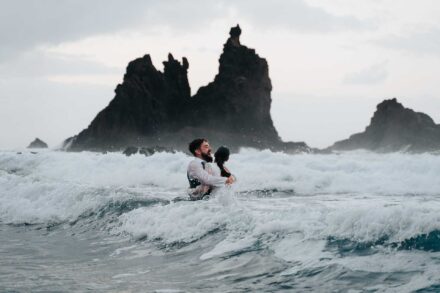 Los novios, abrazados en medio de las olas que rompen, con grandes rocas oscuras detrás