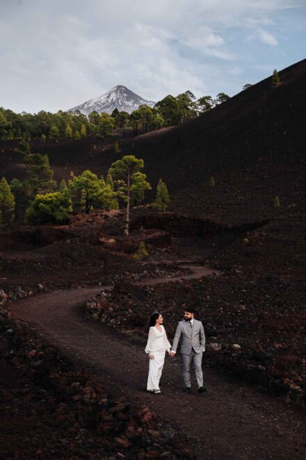 Los novios se miran de la mano en un camino serpenteante de tierra volcánica, con la cumbre nevada del Teide detrás