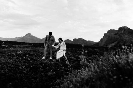 El novio ayuda a la novia a cruzar de la mano un pedregal de lava, con montañas al fondo, en blanco y negro