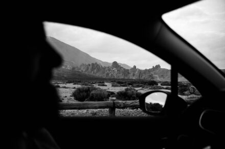 Picos rocosos y matorrales vistos por la ventanilla de un coche, con una silueta a contraluz delante, en blanco y negro