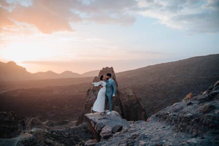 La novia se echa hacia atrás con el brazo extendido mientras el novio la besa sobre una roca al atardecer