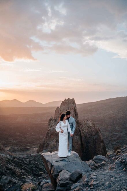Los novios abrazados, cabeza con cabeza, sobre una roca delante de un pitón volcánico y un cielo de atardecer