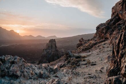 Sendero entre rocas volcánicas y un pitón de piedra, con el sol poniéndose tras las montañas