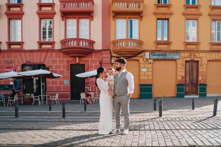 Plano abierto de los novios con un bebé en brazos en una calle adoquinada con raíles de tranvía y fachadas de colores