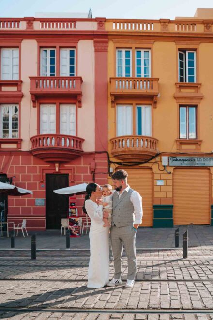 La novia besa a un bebé en brazos del novio, ante fachadas rosa y amarilla con balcones, en Santa Cruz de Tenerife