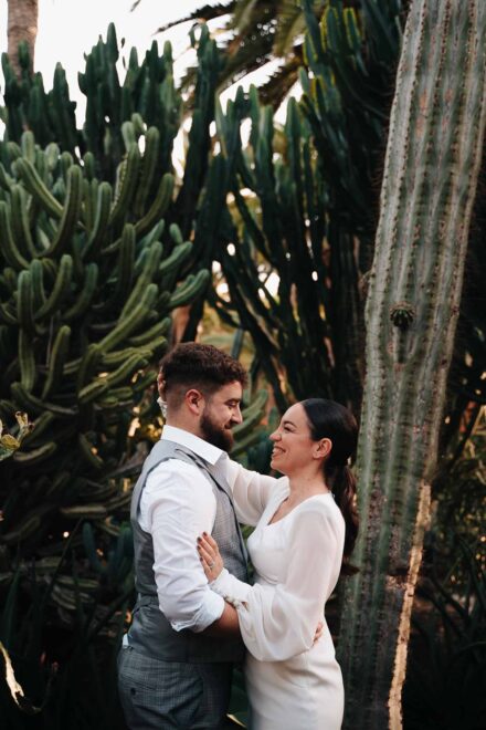 Los novios abrazados se sonríen entre cactus altos y palmeras
