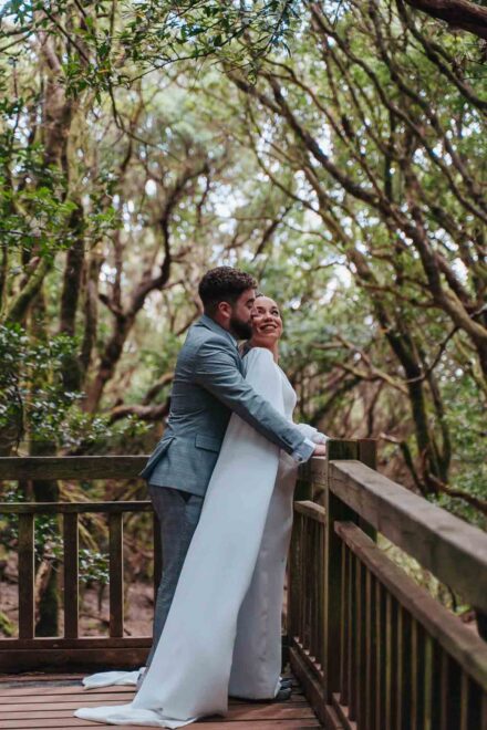 La novia sonríe apoyada en la barandilla de una pasarela del bosque mientras el novio la abraza por detrás