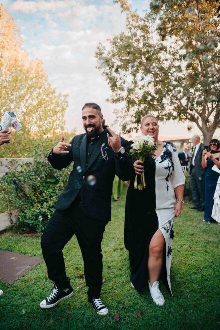 Los novios salen con zapatillas entre pompas de jabón, él haciendo cuernos con las manos y ella con calas