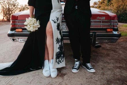Los novios de cintura para abajo ante un coche rojo antiguo: ella, de blanco y negro con zapatillas blancas; él, de negro