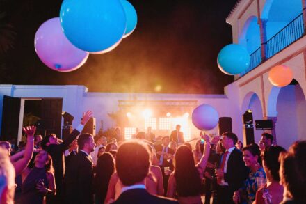 Los invitados juegan de noche con globos gigantes de colores en un patio de arcos blancos, con la banda al fondo