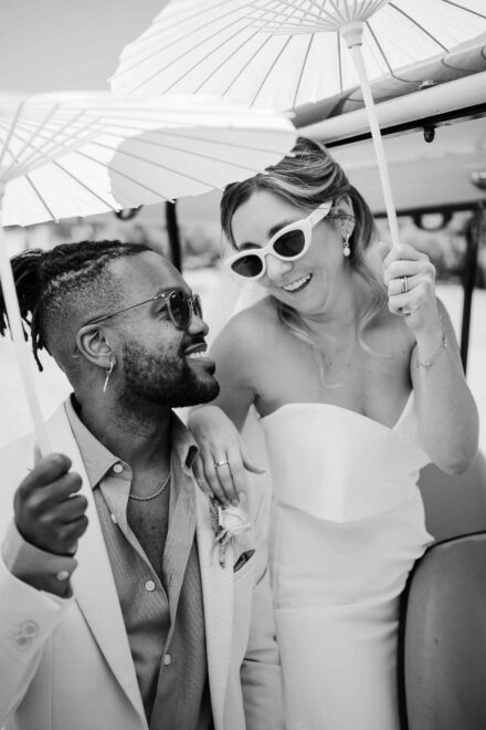 El novio mira sonriente a la novia, que ríe con gafas blancas bajo una sombrilla de papel, en blanco y negro