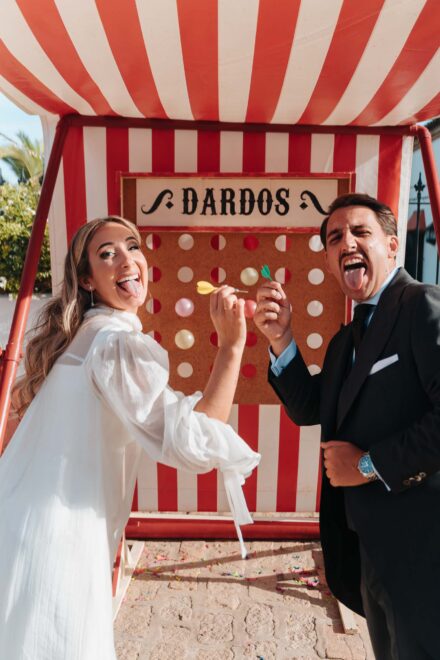 Los novios sacan la lengua mientras lanzan dardos en una caseta de feria de rayas rojas y blancas