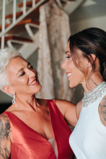 La novia y una mujer de pelo blanco corto y vestido rojo se miran sonriendo cara a cara