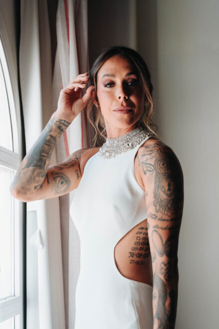 La novia, con collar joya y los brazos tatuados, posa junto a la ventana con una mano en el pelo