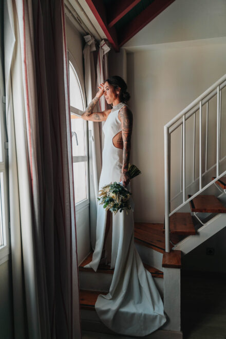 La novia, de cuerpo entero y con el ramo, mira por una ventana en arco al pie de la escalera
