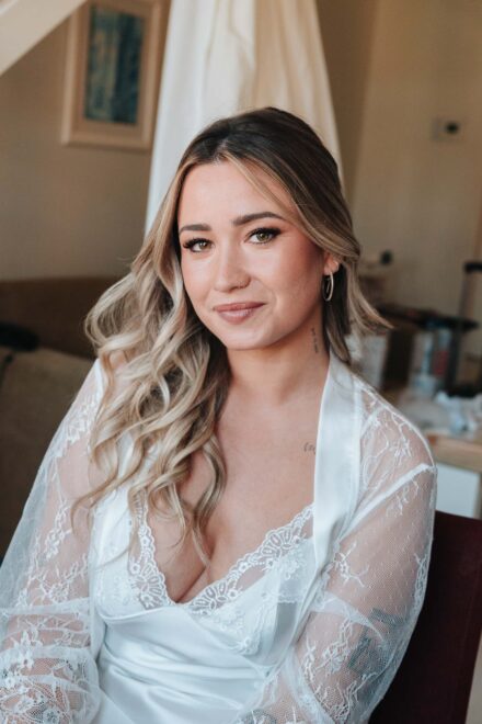 La novia, con melena rubia ondulada y bata de encaje, sonríe a cámara ya maquillada