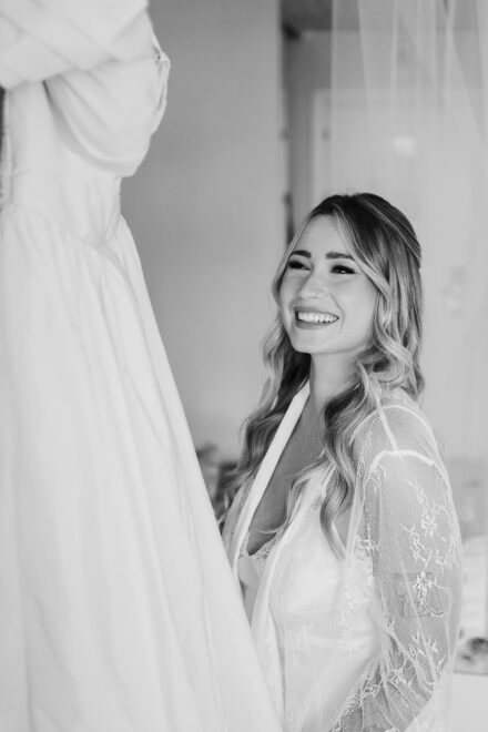 La novia sonríe en bata de encaje mirando su vestido de novia colgado, en blanco y negro
