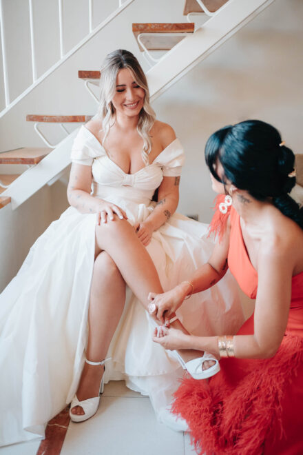 Una invitada de vestido coral con plumas abrocha la sandalia a la novia, sentada en la escalera