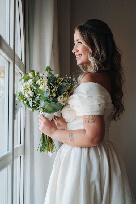 La novia sonríe de perfil junto a la ventana con el ramo y el vestido de hombros caídos