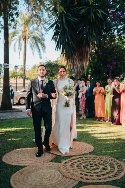 La novia avanza del brazo de un chico por el pasillo de alfombras de esparto, en Finca Al-Yamanah (Sevilla)