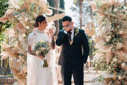 La novia sonríe ante el altar de flores secas mientras el chico que la acompaña se tapa la cara emocionado