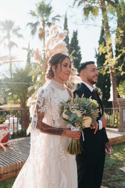 La novia, con capa de encaje y el ramo, espera sonriendo junto al chico que la acompaña, a contraluz