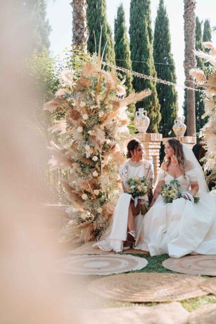 Lola Gallardo y Cristina sentadas junto a un arreglo de pampas durante la ceremonia, en Finca Al-Yamanah (Sevilla)