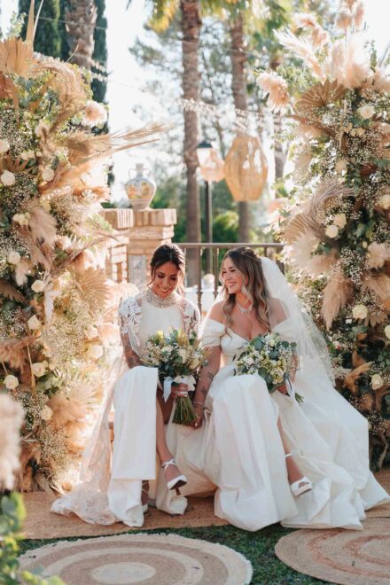 Las novias ríen sentadas entre las dos columnas de pampas y flores, con los ramos en el regazo