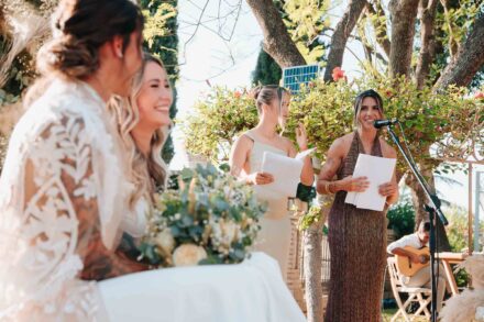 Andrea Medina y Marta Carmona leen unos papeles ante las novias, con un guitarrista detrás, durante la ceremonia
