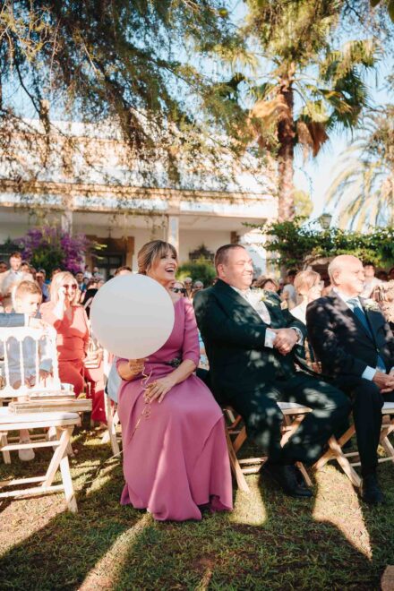 Una invitada de rosa con un globo blanco ríe sentada entre los invitados de la ceremonia