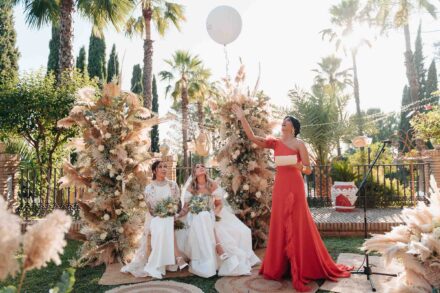 Una invitada de rojo suelta un globo blanco junto a las novias, sentadas entre las pampas de la ceremonia