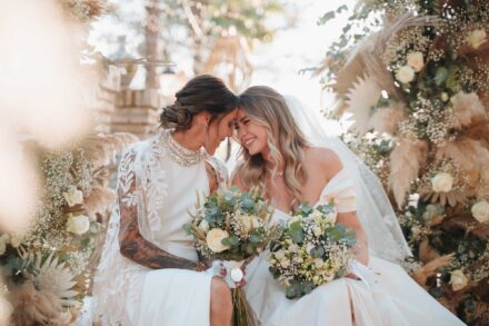 Las novias juntan las frentes y sonríen, sentadas con sus ramos entre las flores de la ceremonia